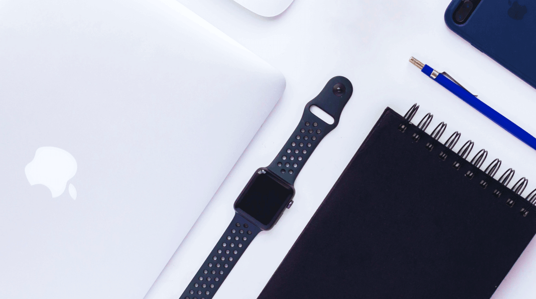 На столі з білим фоном розташовані: срібний ноутбук Apple, чорний смарт-годинник, темний блокнот з спіраллю, синій ручка і смартфон чорного кольору.