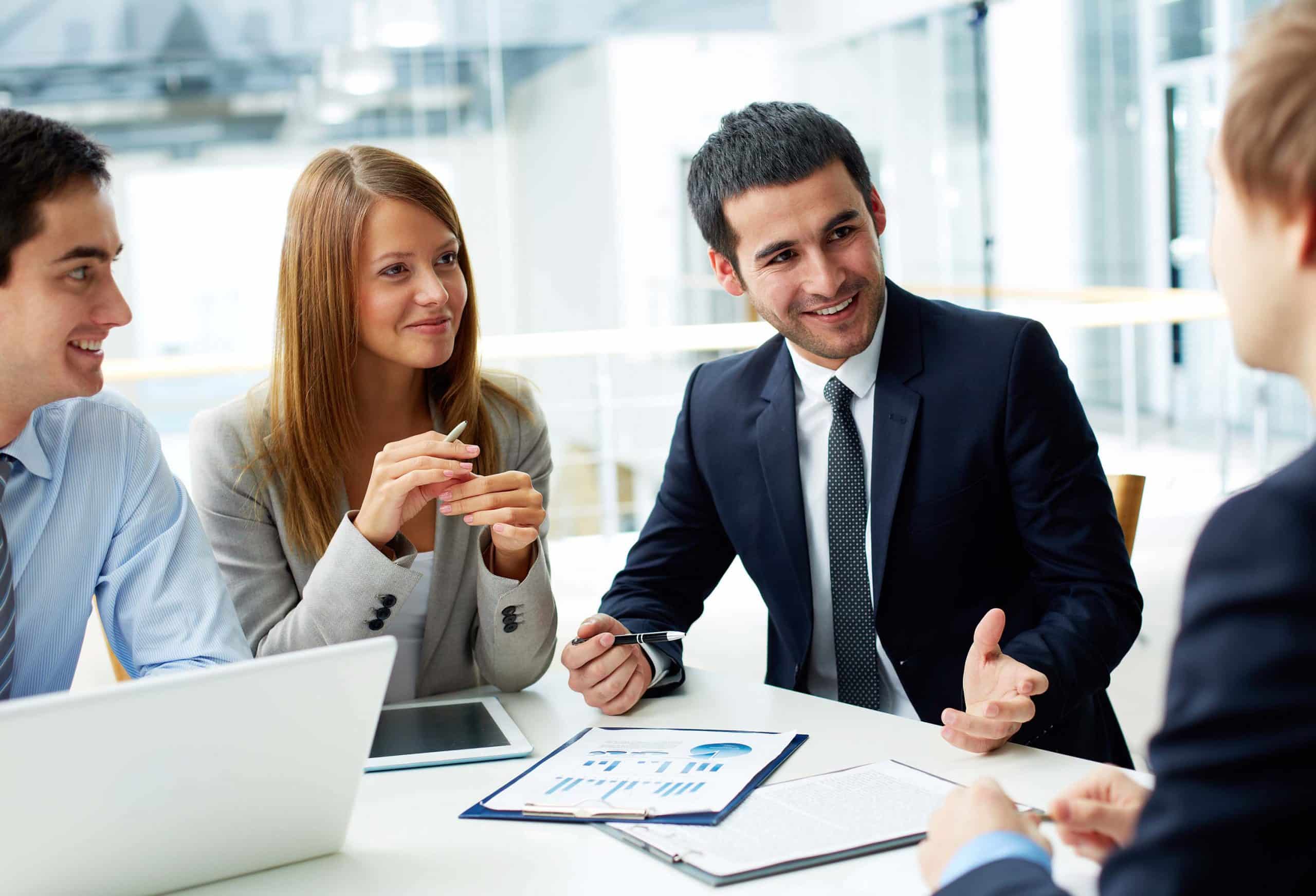 Четверо молодих колег у бізнес-вбранні обговорюють ідеї під час зустрічі в сучасному офісі. На столі лежать документи і планшет, а ведучий презентує свої думки, усміхаючись.