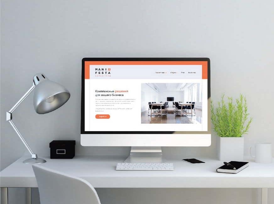 Сучасний робочий стіл з комп'ютером, на екрані якого веб-сайт на тему офісних приміщень. Лампа, чорний блокнот і зелений рослинний елемент доповнюють простір.