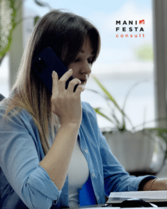 Жінка розмовляє по телефону, тримаючи слухавку біля вуха, сидячи за столом з рослинами на фоні. Вона одягнена у блакитну сорочку та має розпущене волосся. На столі видно канцелярські приладдя.